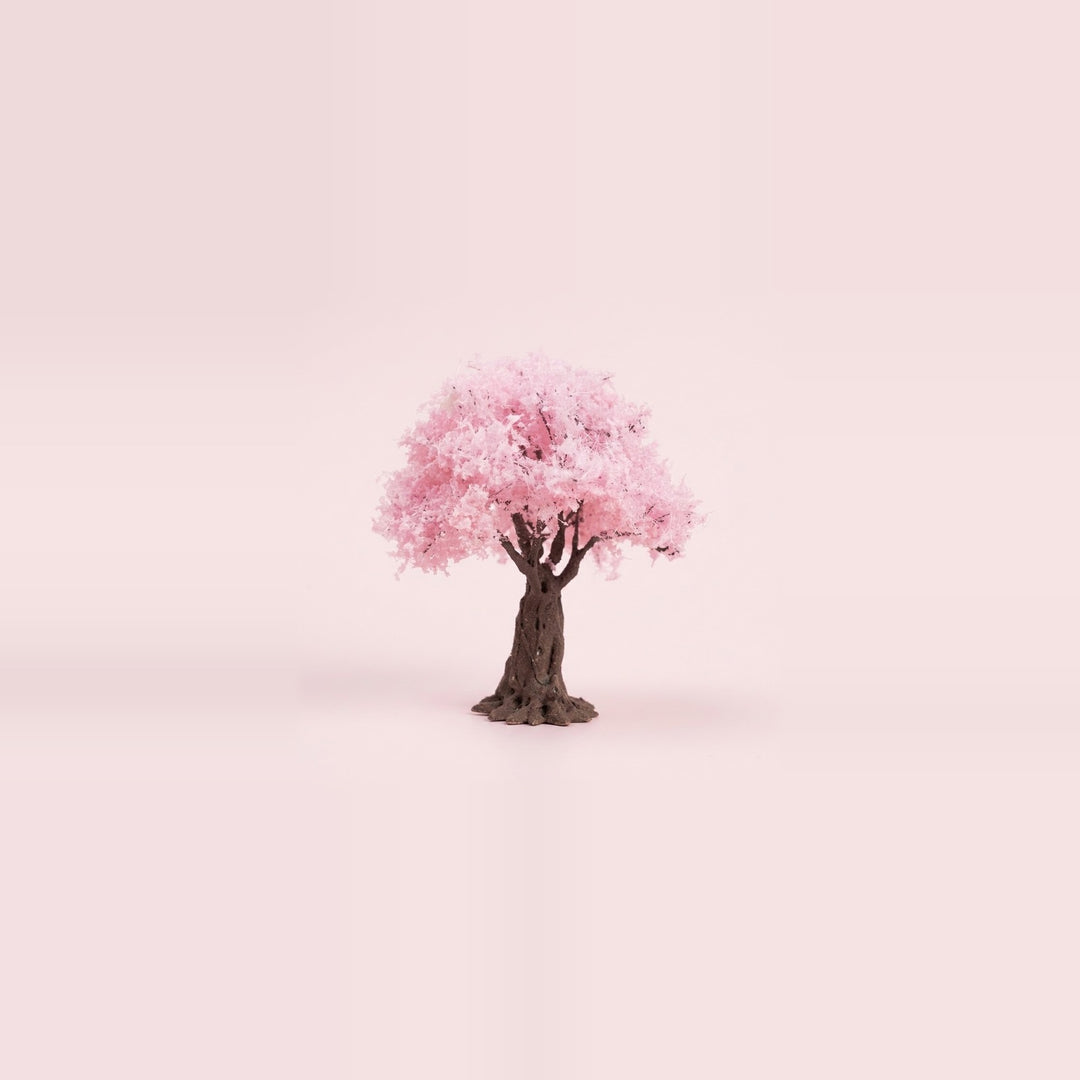 A miniature model blossom tree with pink foliage and a dark brown trunk on a soft pink studio background, 3.9 inches (10 cm) tall.