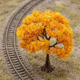 An orange miniature tree with a brown trunk stands beside curved model railway tracks on gravelly terrain, viewed from above.