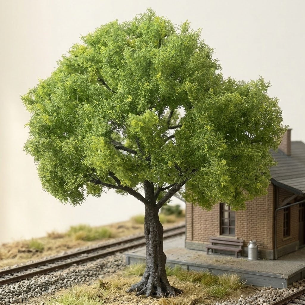A light green miniature tree with a dark trunk stands beside a model railway station, tracks, and grassy gravel terrain.