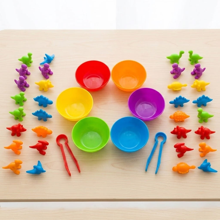 Sorting toy set with colourful dinosaur pieces surrounding six rainbow bowls and red and blue tweezers on a wooden table.