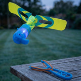 A blue plastic slingshot helicopter with yellow and teal rotor blades glows above a wooden table holding its launcher outdoors.