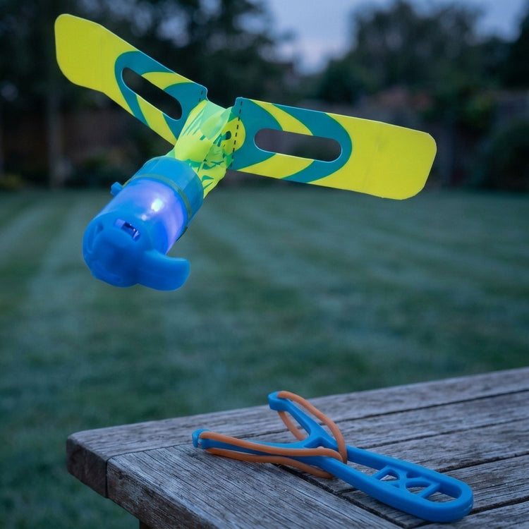 A blue plastic slingshot helicopter with yellow and teal rotor blades glows above a wooden table holding its launcher outdoors.