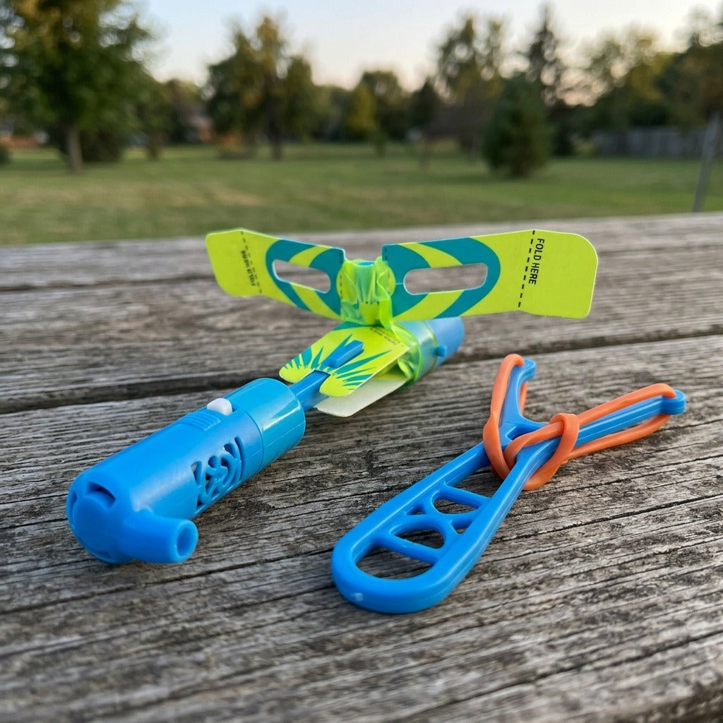 A blue plastic slingshot helicopter with yellow-green patterned wings rests beside its blue launcher and orange elastic on a weathered picnic table.