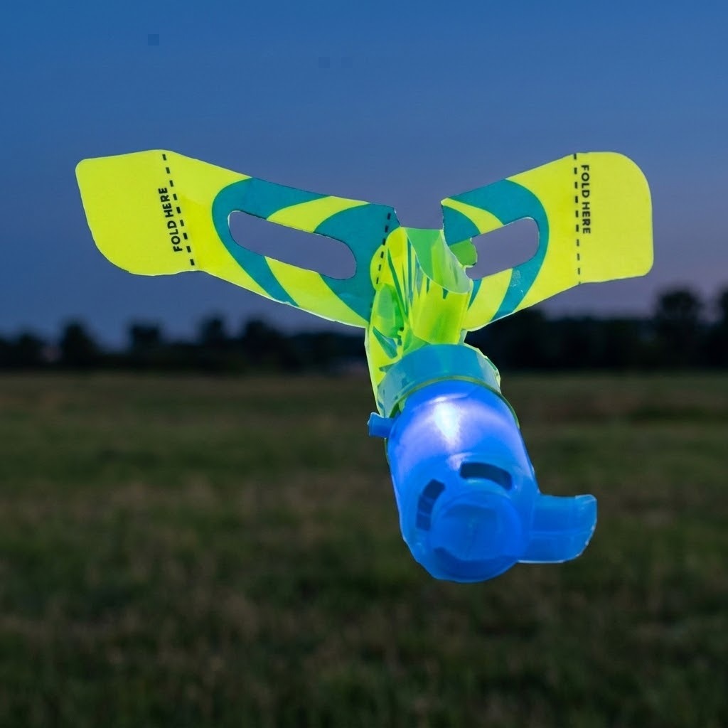 A blue plastic slingshot helicopter glows above a lawn at dusk, with yellow and teal rotor blades marked FOLD HERE.