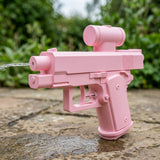 A pink toy water gun shaped like a pistol with a cylindrical sight, shown outdoors on a wet stone surface with blurred green foliage in the background. A stream of water is visible shooting from the front nozzle.