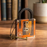 A square wooden puzzle lock with a black shackle and a small brass key resting in front, placed on a wooden surface with books and a plant blurred in the background.