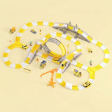 A children's toy train track set arranged on a pale beige background. The set includes a yellow and white looped track, a curved track section, a bridge, an orange crane, small toy cars, and tiny construction-themed accessories.