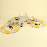 A toy construction-themed winding track set with yellow and white curved rails, a gray suspension bridge, small toy vehicles including a yellow car, excavator, and dump truck, and a small palm tree, photographed on a plain warm beige background.