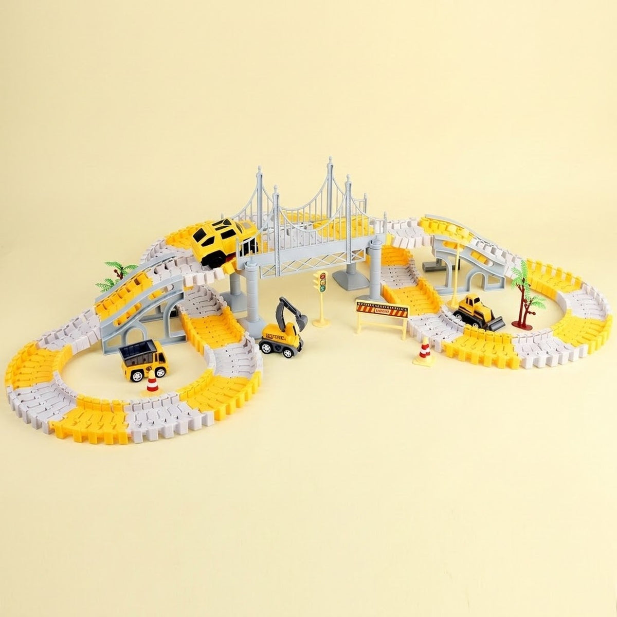 A toy construction-themed winding track set with yellow and white curved rails, a gray suspension bridge, small toy vehicles including a yellow car, excavator, and dump truck, and a small palm tree, photographed on a plain warm beige background.