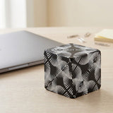 A closed black and white geometric puzzle cube rests on a light wooden desk beside a closed silver laptop.