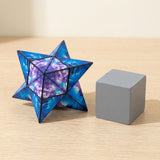 Blue and purple galaxy-patterned puzzle cube folded into a pointed star beside a plain grey cube on a light wooden table.