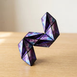 Purple and blue galaxy-patterned puzzle cube folded into a twisting upright shape on a light wooden table against a neutral background.