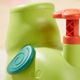 Close-up of the green plastic elephant disc launcher shows its teal spiral ear and raised orange pedal against a cream background.