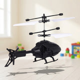 A black remote control helicopter with stacked white rotor blades hovers above a pale wooden floor with colorful blocks behind it.