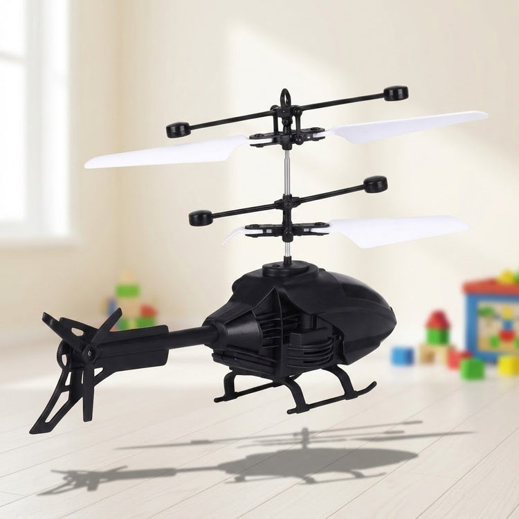 A black remote control helicopter with stacked white rotor blades hovers above a pale wooden floor with colorful blocks behind it.