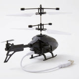 A black remote control helicopter with white rotor blades sits on a white surface with a white USB charging cable connected.