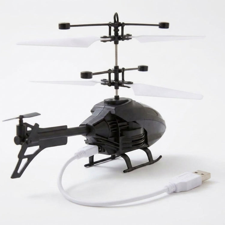 A black remote control helicopter with white rotor blades sits on a white surface with a white USB charging cable connected.