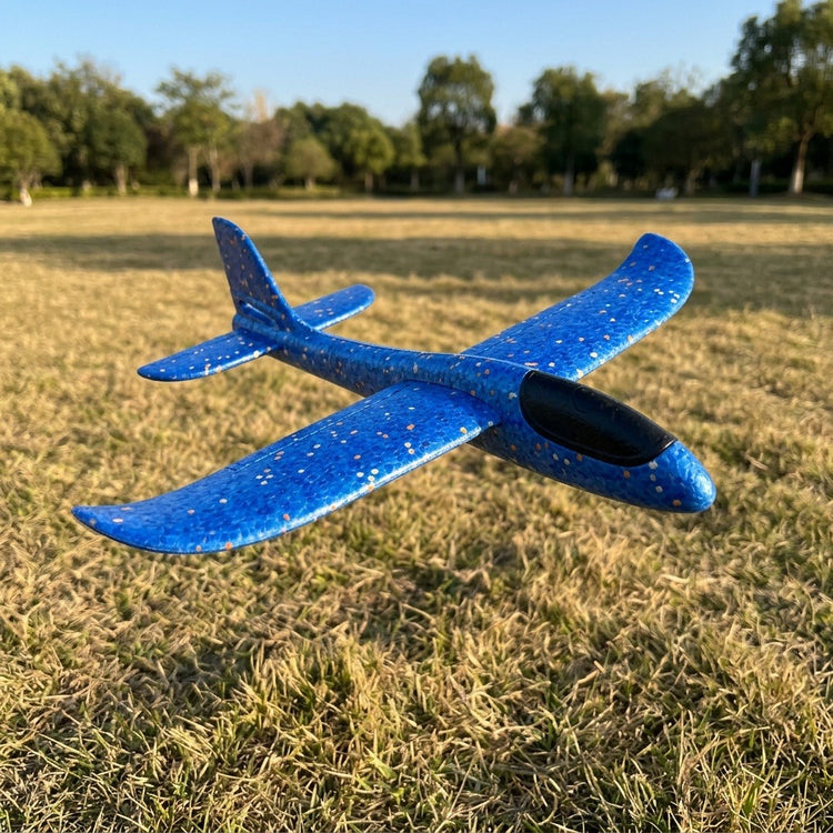 A blue speckled foam toy glider with a black cockpit appears above dry grass, with trees and blue sky behind it.