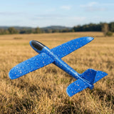 A blue speckled foam toy glider with a black cockpit is viewed from above against a blurred dry grass field.