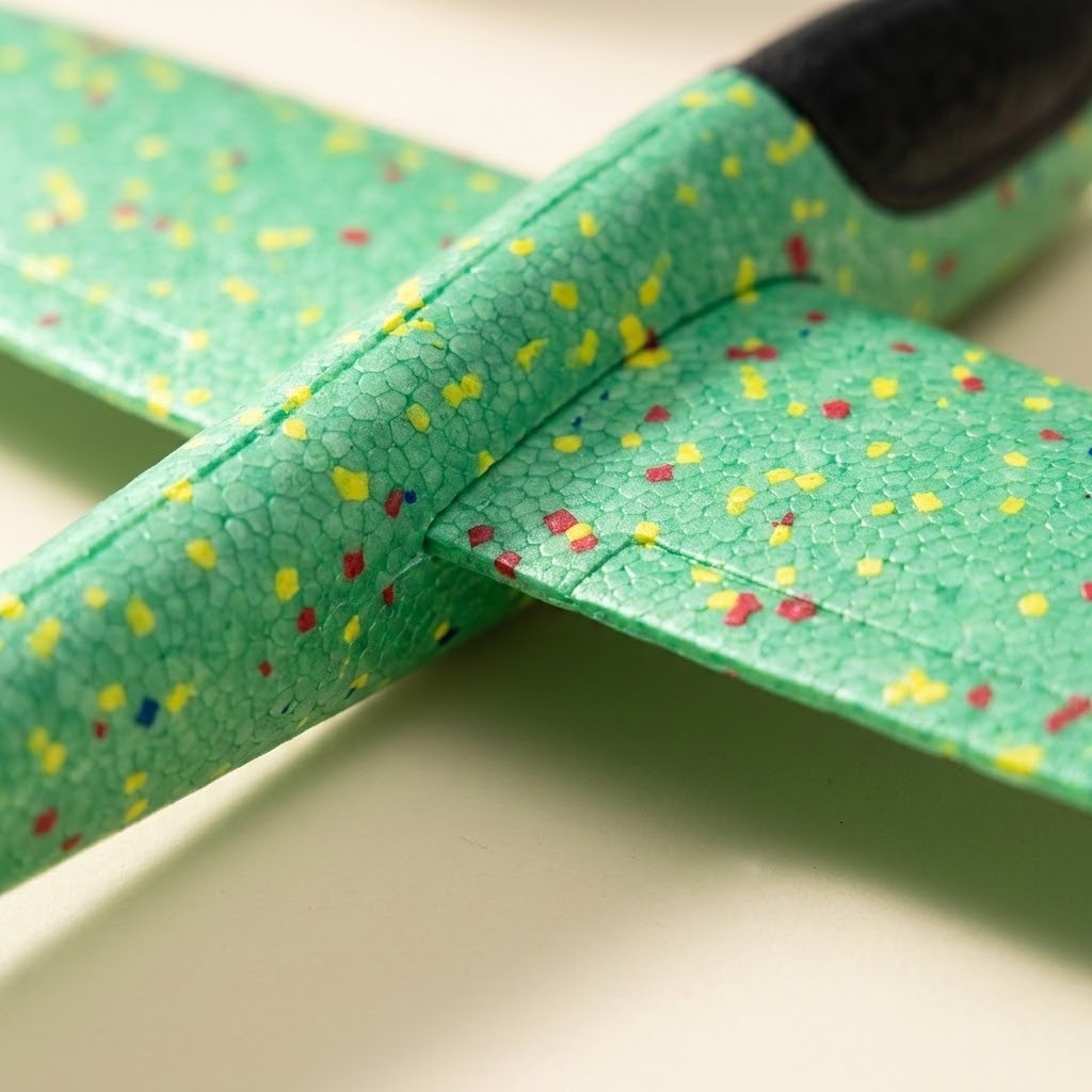 A close-up of a green foam toy glider shows its pebbled texture, multicolored speckles, and wing joint against a pale background.