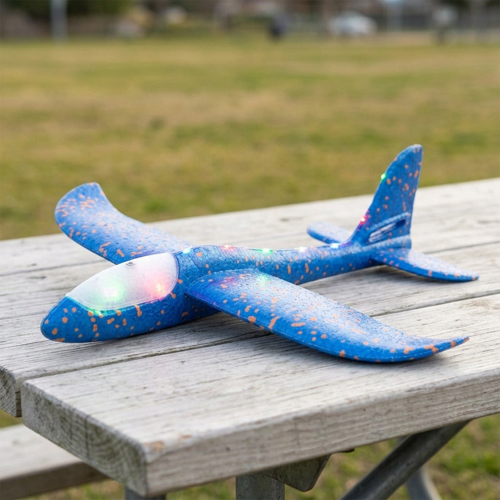 A blue speckled foam toy glider with a glowing cockpit and multicolored lights rests on a weathered wooden picnic table outdoors.