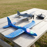 A blue foam toy glider with glowing multicoloured LEDs rests on a wooden outdoor table beside a grey propeller airplane and controller.