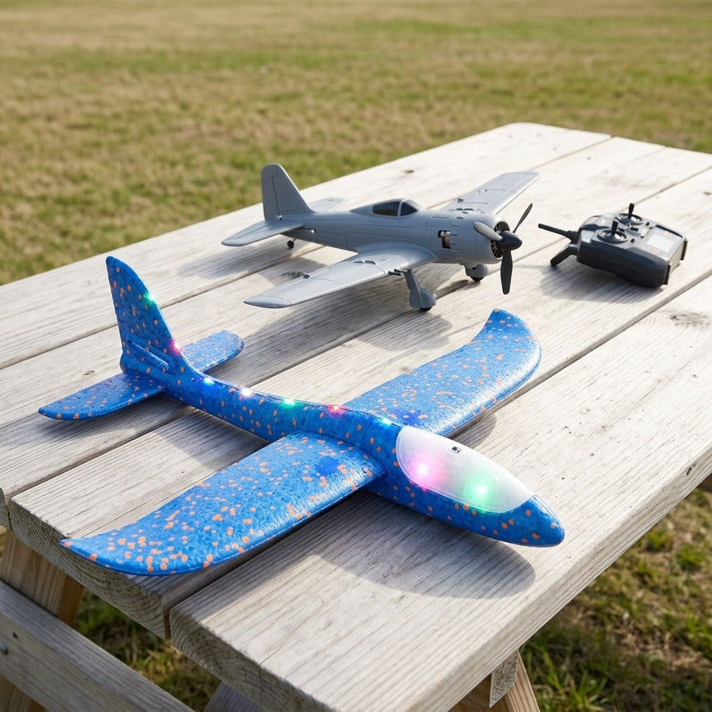 A blue foam toy glider with glowing multicoloured LEDs rests on a wooden outdoor table beside a grey propeller airplane and controller.