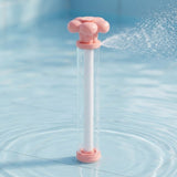 A pink and clear water gun stands upright in shallow blue pool water, spraying a fan of water sideways.