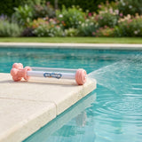 A pink and clear plastic water gun rests on a pale pool edge, spraying a fan of water over turquoise water.