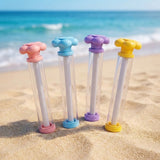 Four clear water guns with pink, blue, purple, and yellow flower-shaped handles stand on sand with ocean waves behind them.