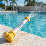 A yellow and clear water gun rests diagonally on a sunlit pool edge, spraying water with palm trees in the background.