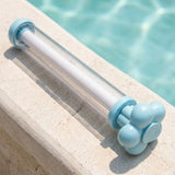 A clear water gun with light blue ends and a white inner tube rests on beige stone beside turquoise pool water.