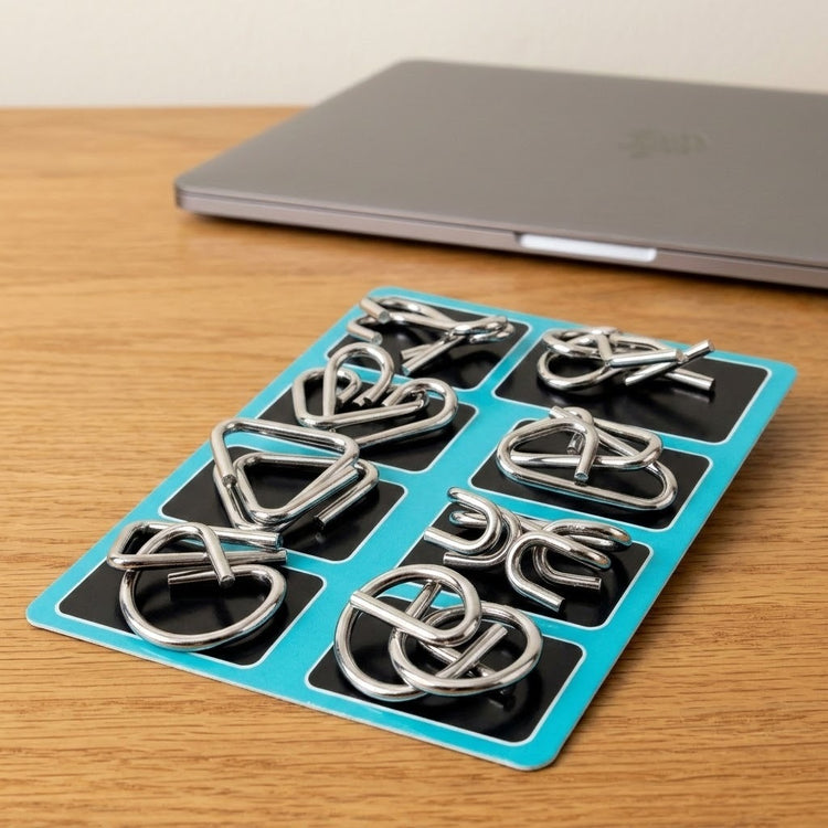 Eight-piece silver-colored wire brain teaser set on a turquoise backing card beside a closed laptop on a wooden tabletop.