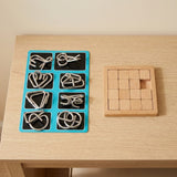 Silver metal wire brain teaser set on a turquoise backing beside a wooden sliding puzzle on a light wooden table.