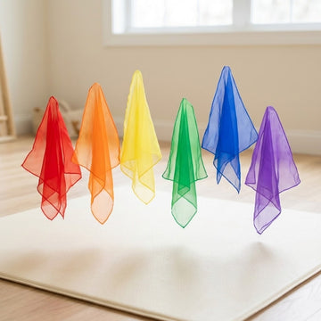 Six sheer chiffon juggling scarves in rainbow colours float in a row above a cream mat in a bright room.