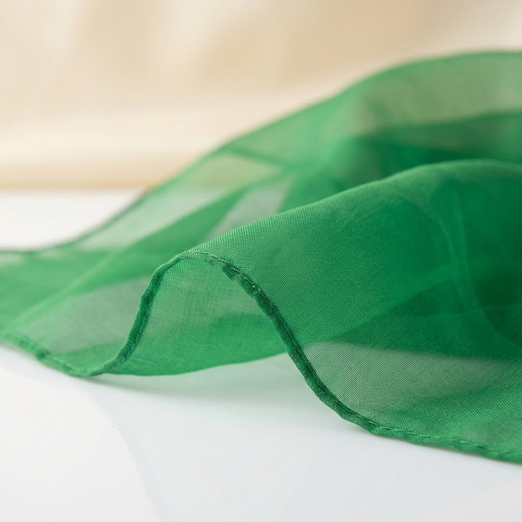 A close-up of a sheer green fabric scarf or sash draped elegantly, showing its lightweight texture and fine weave against a soft, light beige and white background.