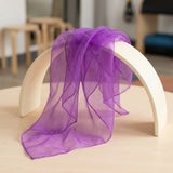 A sheer purple scarf draped over a cream-colored arched stand on a light wood table, with a softly blurred indoor background.