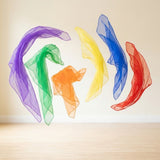 Six sheer chiffon juggling scarves in purple, green, orange, yellow, blue, and red float above a wooden surface against a cream background.