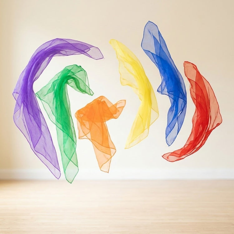Six sheer chiffon juggling scarves in purple, green, orange, yellow, blue, and red float above a wooden surface against a cream background.