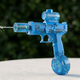 A translucent blue electric water gun with a round lower tank sprays water on a pale surface against blurred greenery.