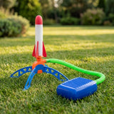 A plastic toy rocket launcher set on a green grass lawn, featuring a white rocket with red fins and nose cone, a blue support stand, a green hose, and a blue foot pump.