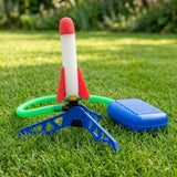 Rocket launcher toy on a sunny lawn with an upright red-and-white rocket, blue stand and foot pump, and looped green hose.