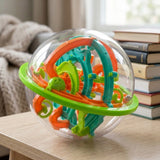 A clear spherical maze puzzle with orange, teal and lime-green plastic tracks rests on a wooden table beside stacked books.