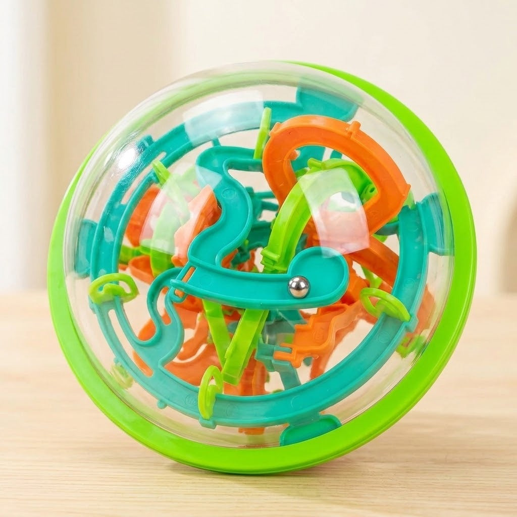 A clear plastic maze puzzle with a green rim, teal and orange tracks, and a visible steel ball stands on a pale wooden surface.