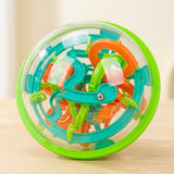 A clear plastic maze puzzle with a green rim, teal and orange tracks, and a visible steel ball stands on a pale wooden surface.