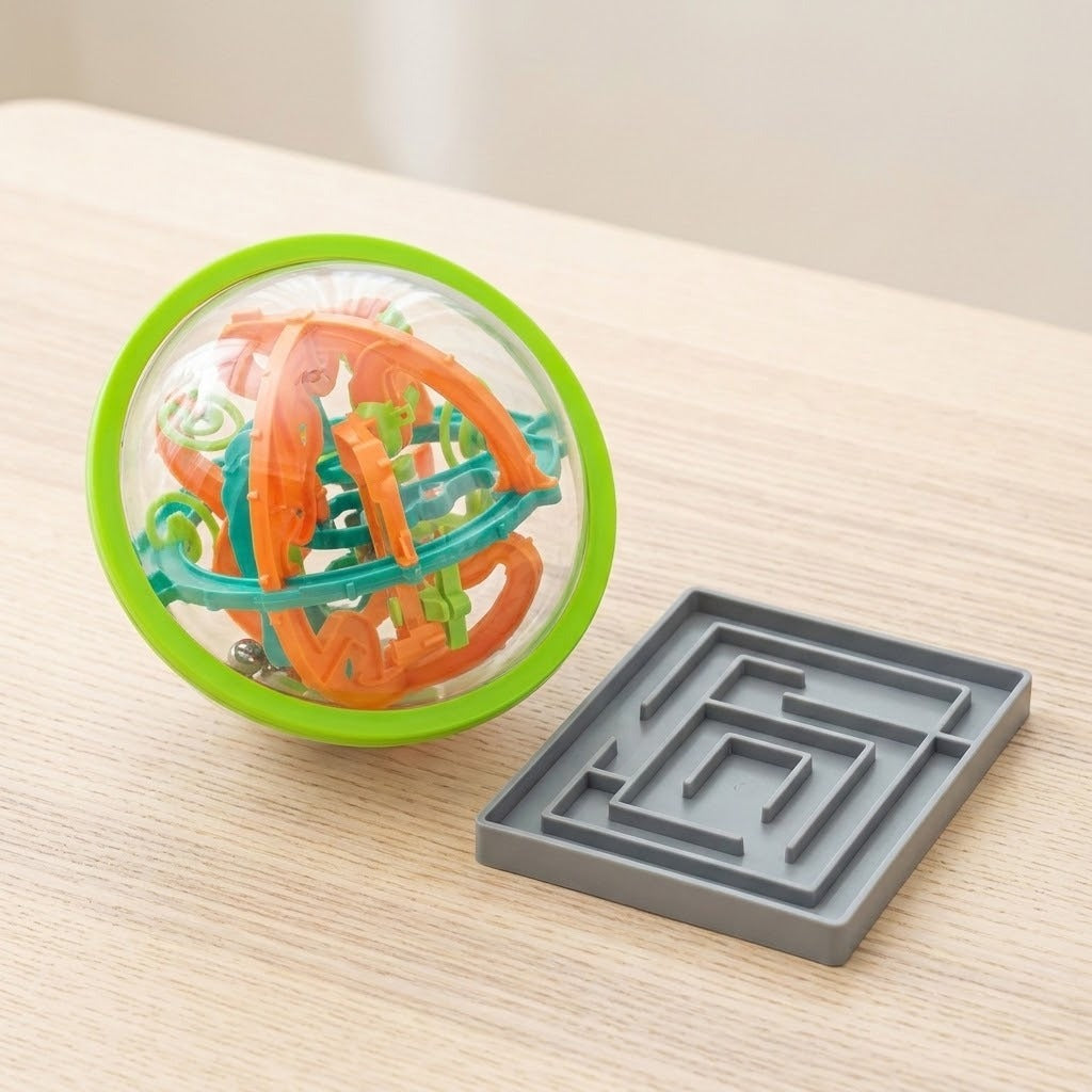 A clear spherical maze puzzle with a green rim and orange and teal tracks sits beside a flat gray maze on a wooden table.
