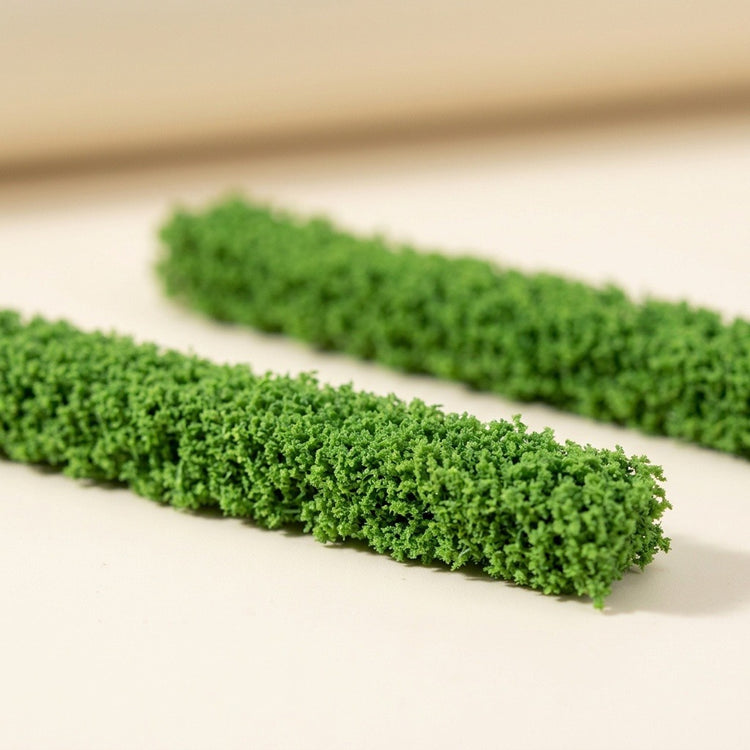 Two green model hedges rest on a cream surface, with the foreground strip showing detailed foliage texture.