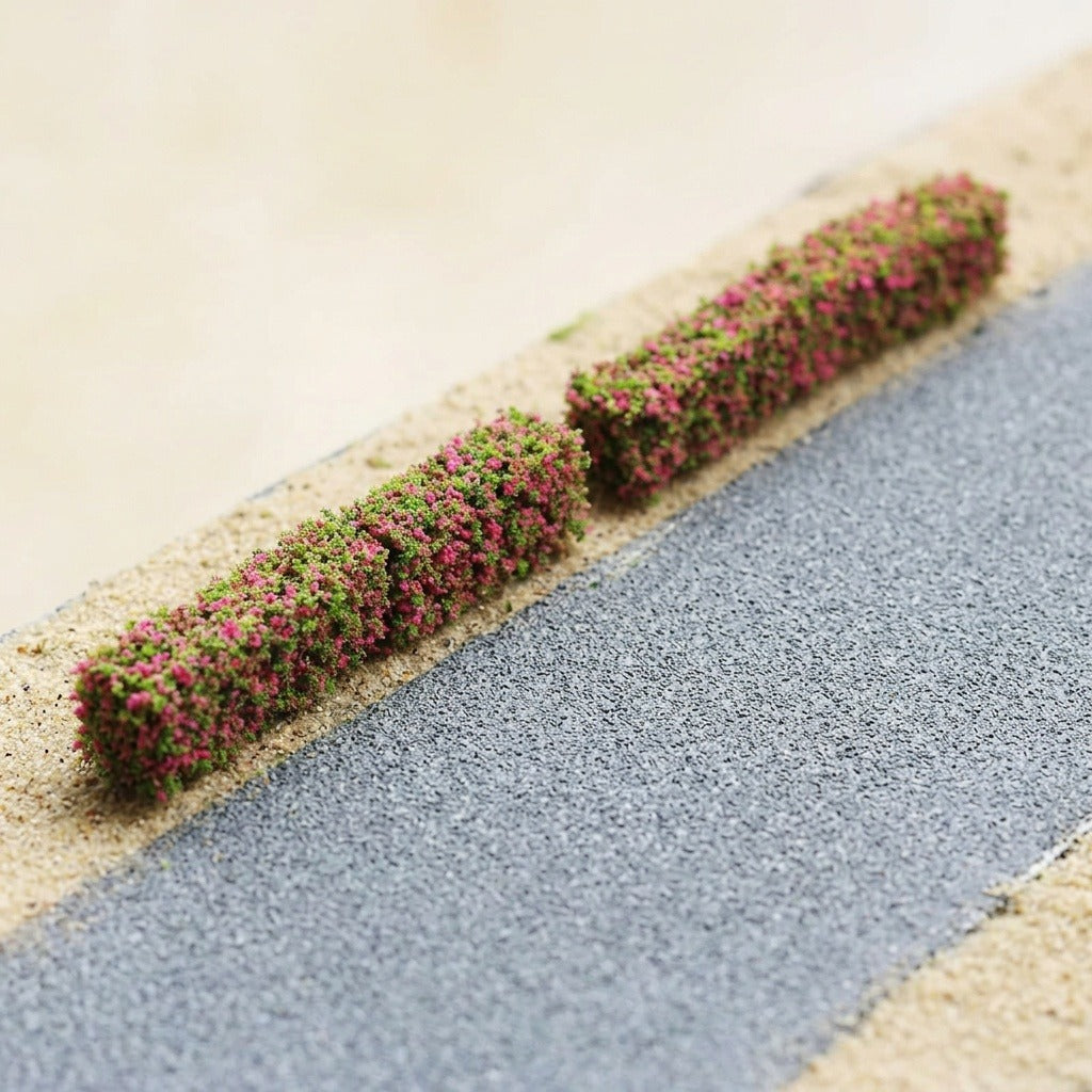 Two pink and green miniature hedge strips sit end to end beside a gray model road on sandy terrain.