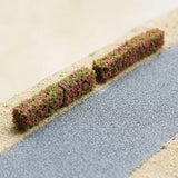 Two pink and green miniature hedge strips sit end to end beside a gray model road on sandy terrain.