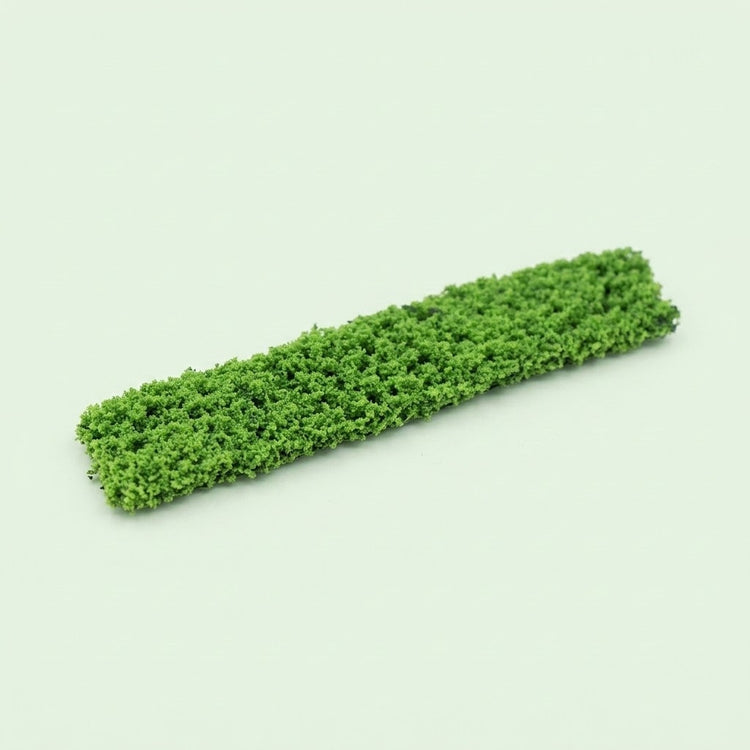 A miniature model hedge with dense green textured foliage lies diagonally on a pale green background.
